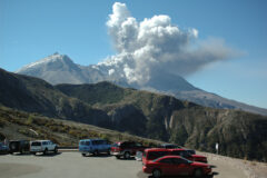 volcano venting steam and ash under clear daytime sky and cars in the parking lot